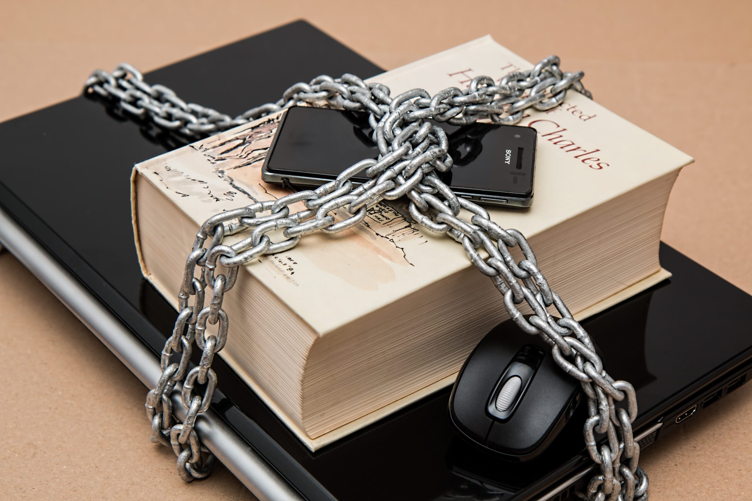 Stack of laptop, hardcover book and mobile phone all tied and locked together by a silver chains.